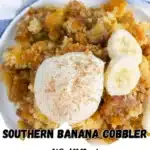 A serving of warm Southern Banana Cobbler topped with a scoop of vanilla ice cream and banana slices, presented on a white plate with a blue checkered napkin in the background.