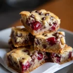 A close-up view of stacked chocolate chip cherry bars on a white plate, showcasing their golden edges, gooey cherry centers, and scattered chocolate chips.