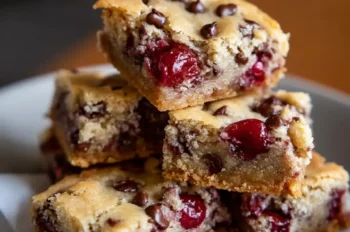 A close-up view of stacked chocolate chip cherry bars on a white plate, showcasing their golden edges, gooey cherry centers, and scattered chocolate chips.