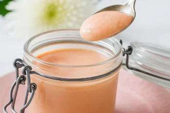 A spoon lifting creamy pink rhubarb curd from a glass jar, with a white flower in the background.