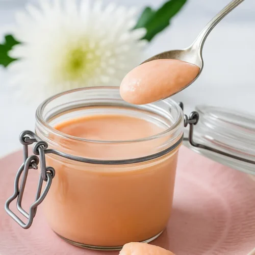 A spoon lifting creamy pink rhubarb curd from a glass jar, with a white flower in the background.