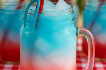 Layered red, white, and blue kid-friendly patriotic punch served in a mason jar with ice and red licorice candy straw tied with blue candy lace