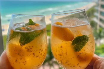 Close-up of a sparkling peach and apricot drink with visible fruit slices and bubbles, served in a clear glass with lemon and mint.