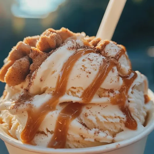 Churro Cinnamon Delight Ice Cream served in a white cup with a spoon, drizzled with caramel and topped with crumbled churro pieces against a sunny outdoor backdrop.