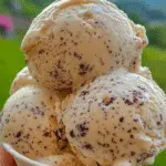 Close-up of creamy cottage cheese ice cream with chocolate flecks, served in a white paper cup and held outdoors with a mountain landscape in the background.