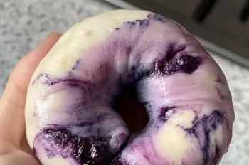 A hand holding a freshly shaped marbled blueberry bagel with visible blueberry swirls and deep purple spots against soft white dough.