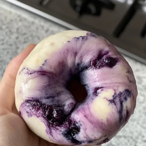 A hand holding a freshly shaped marbled blueberry bagel with visible blueberry swirls and deep purple spots against soft white dough.