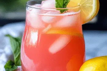 Glass of vibrant pink rhubarb lemonade over ice, garnished with fresh mint and a lemon slice, on a marble surface with a lemon and white napkin beside it.