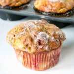 Close-up of a moist rhubarb muffin in a pink-striped paper liner, topped with cinnamon sugar, with a pan of muffins in the background.