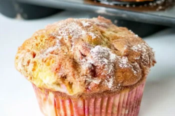 Close-up of a moist rhubarb muffin in a pink-striped paper liner, topped with cinnamon sugar, with a pan of muffins in the background.