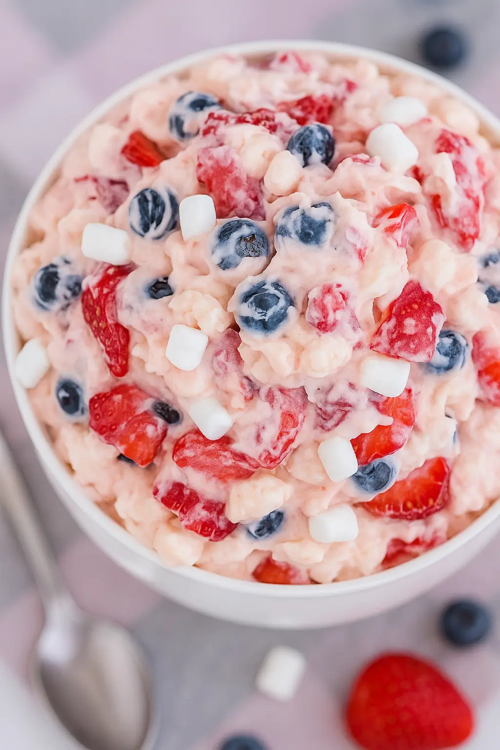 Red White and Blue Cheesecake Salad being mixed in a glass bowl with a red spatula, showing strawberries, blueberries, and marshmallows.