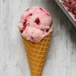Two scoops of rhubarb berry no churn ice cream in a waffle cone on a white wooden background with ice cream container nearby