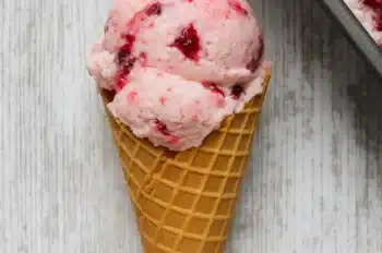 Two scoops of rhubarb berry no churn ice cream in a waffle cone on a white wooden background with ice cream container nearby