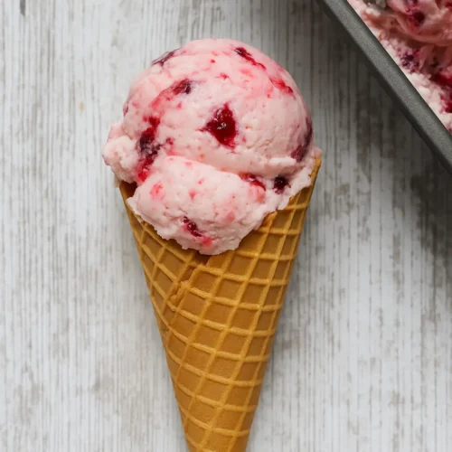 Two scoops of rhubarb berry no churn ice cream in a waffle cone on a white wooden background with ice cream container nearby