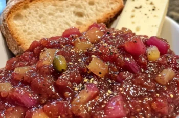 Spiced rhubarb chutney served in a white bowl, garnished with black pepper, accompanied by crusty bread slices and a wedge of cheese on a stone countertop.