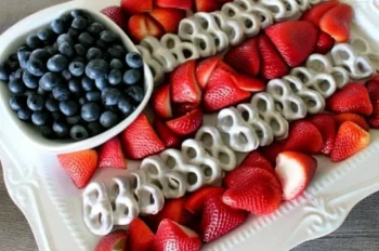 Fourth of July fruit platter arranged in the shape of an American flag using fresh blueberries in a small white bowl for the stars section, with alternating rows of sliced strawberries and yogurt-covered pretzels forming the stripes, all displayed on an ornate white serving tray