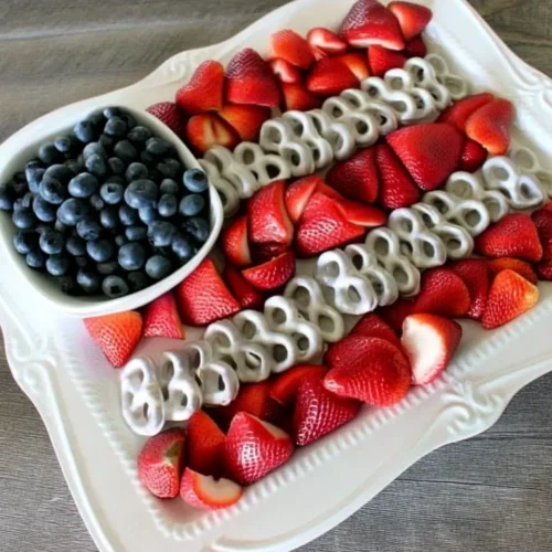 Fourth of July fruit platter arranged in the shape of an American flag using fresh blueberries in a small white bowl for the stars section, with alternating rows of sliced strawberries and yogurt-covered pretzels forming the stripes, all displayed on an ornate white serving tray