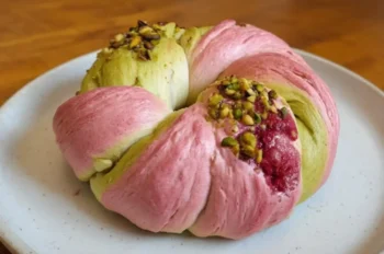 A vibrant raspberry and pistachio sourdough bun with pink, green, and white dough sections, topped with chopped pistachios and whole raspberries, served on a white plate on a wooden table