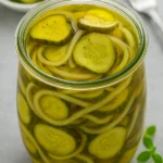 Glass jar filled with bread and butter pickles and sliced onions in golden brine on a gray background