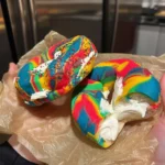 Two vibrant rainbow bagels filled with whipped cream cheese, held in a person's hands over brown parchment paper