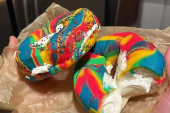 Two vibrant rainbow bagels filled with whipped cream cheese, held in a person's hands over brown parchment paper