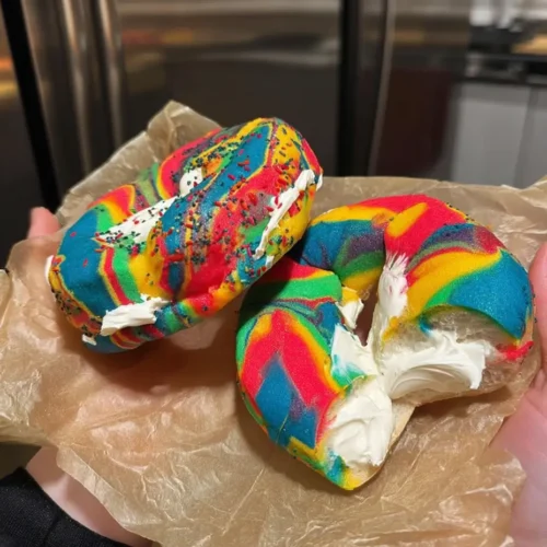 Two vibrant rainbow bagels filled with whipped cream cheese, held in a person's hands over brown parchment paper