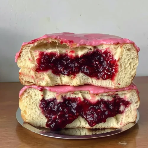Front view of two brioche donuts stacked on a small silver tray, sliced to reveal a thick, juicy rhubarb-strawberry jam filling with bright pink glaze dripping slightly over the edges.