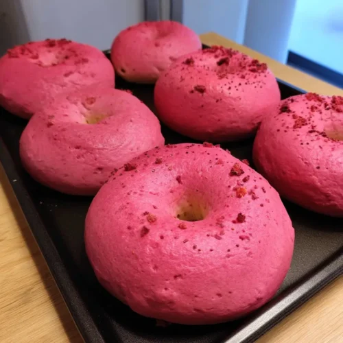 Close-up of six bright pink raspberry sourdough bagels on a black baking tray, showing their smooth, fluffy texture and rich color.