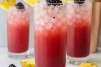 Three tall glasses of pineapple ginger beer mocktail with crushed ice, garnished with pineapple wedges, blackberries, and green leaves, surrounded by scattered ice and fresh berries.