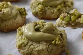 Buttery pistachio cookies with a thumbprint center filled with pistachio cream and garnished with crushed pistachios, arranged in rows for cooling