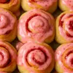 Freshly baked strawberry cinnamon rolls topped with glossy pink strawberry glaze, arranged closely in a baking pan.
