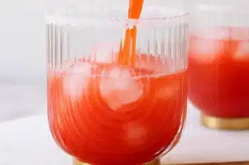 Strawberry lime mocktail margarita being poured from a cocktail shaker into a glass with ice and a salt rim, with additional glasses in the background on a white surface.