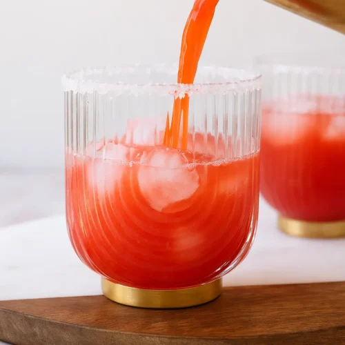 Strawberry lime mocktail margarita being poured from a cocktail shaker into a glass with ice and a salt rim, with additional glasses in the background on a white surface.