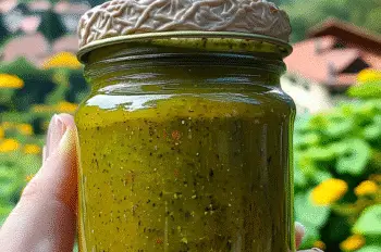 Rustic glass jar filled with zesty homemade dill pickle hot sauce, held outdoors against a picturesque alpine village setting – great for summer grilling