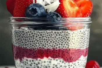 Glass jar of chia pudding layered with strawberries, blueberries, and raspberries, topped with coconut flakes and mint leaves on a marble surface