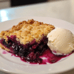 Close-up of blackberry cobbler with juicy berry filling and crisp golden topping, served with a scoop of vanilla ice cream on a square white plate