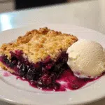 Close-up of blackberry cobbler with juicy berry filling and crisp golden topping, served with a scoop of vanilla ice cream on a square white plate