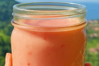 Hand holding a mason jar of pink rhubarb vinaigrette in front of a scenic landscape, overlaid with text instructions for making the vinaigrette.