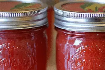 Front-focused view of a mason jar containing rhubarb jam, with other sealed jars and a label reading "Jamaican Rhubarb" visible in the softly blurred background.